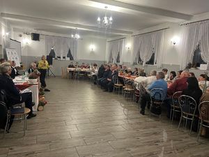 Na zdjęciu widać pomieszczenie sali, wzdłuż której siedzą przy stołach osoby.