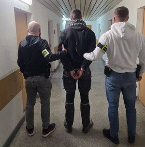 Na korytarzu komendy nieumundurowani policjanci prowadzą zatrzymanego mężczyznę, który ma założone kajdanki.