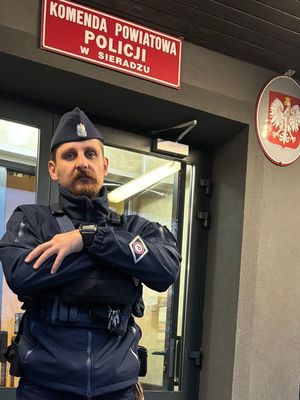 Na zdjęciu widać umundurowanego policjanta, który stoi przed budynkiem Komendy Powiatowej Policji w Sieradzu.