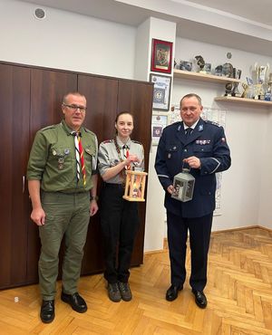 Na zdjęciu z prawej strony znajduję się funkcjonariusz policji, z lewej strony harcerze. Policjant jak i również pani z harcerstwa trzymają                 w dłoniach zapalone lampiony.