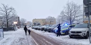 Na parkingu stoją pojazdy, wśród nich  stoi radiowóz z włączonymi sygnałami  świetlnymi, przed nim umundurowana policjantka.