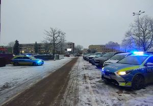 Na parkingu stoją pojazdy, wśród nich  stoją radiowozy z włączonymi sygnałami  świetlnymi.