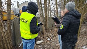 Czynności na miejscu znalezienia truchła psa, stoi nieumundurowani policjant w kamizelce odblaskowej oraz nieumundurowani policjanci z opaskami na ramieniu.
