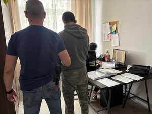 W pomieszczeniu służbowym policjant prowadzi  zatrzymanego mężczyznę, przy biurku siedzi policjantka z opaska na ramieniu  z napisem Policja.