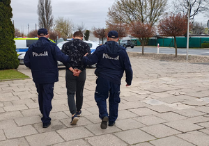 Na parkingu policjanci prowadzą zatrzymanego mężczyznę.