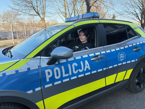 Przed budynkiem komendy na tle oznakowywanego radiowozu stoi umundurowana policjantka.