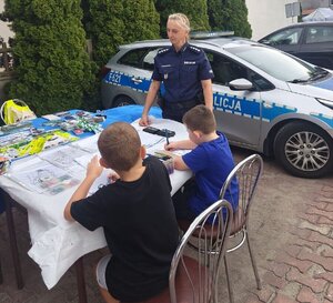 Na zdjęciu widać policjantkę, która stoi przy stole przy którym siedzi dwoje dzieci.