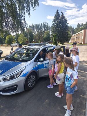 Na zdjęciu widać policjantkę siedząca w radiowozie, drzwi do radiowozu są pootwierane. Przy radiowozie stoją dzieci.