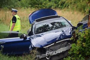 Na zdjęciu widać stojący uszkodzony pojazd koloru niebieskiego, który ma pootwierane boczne drzwi. Za pojazdem tyłem stoi policjant.