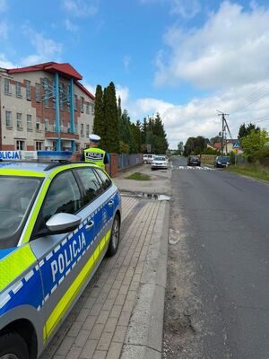 Na zdjęciu widoczny jest radiowóz policyjny za którym stroi funkcjonariusz policji twarzą skierowaną w kierunku budynku, który znajduję się z lewej strony pojazdu.